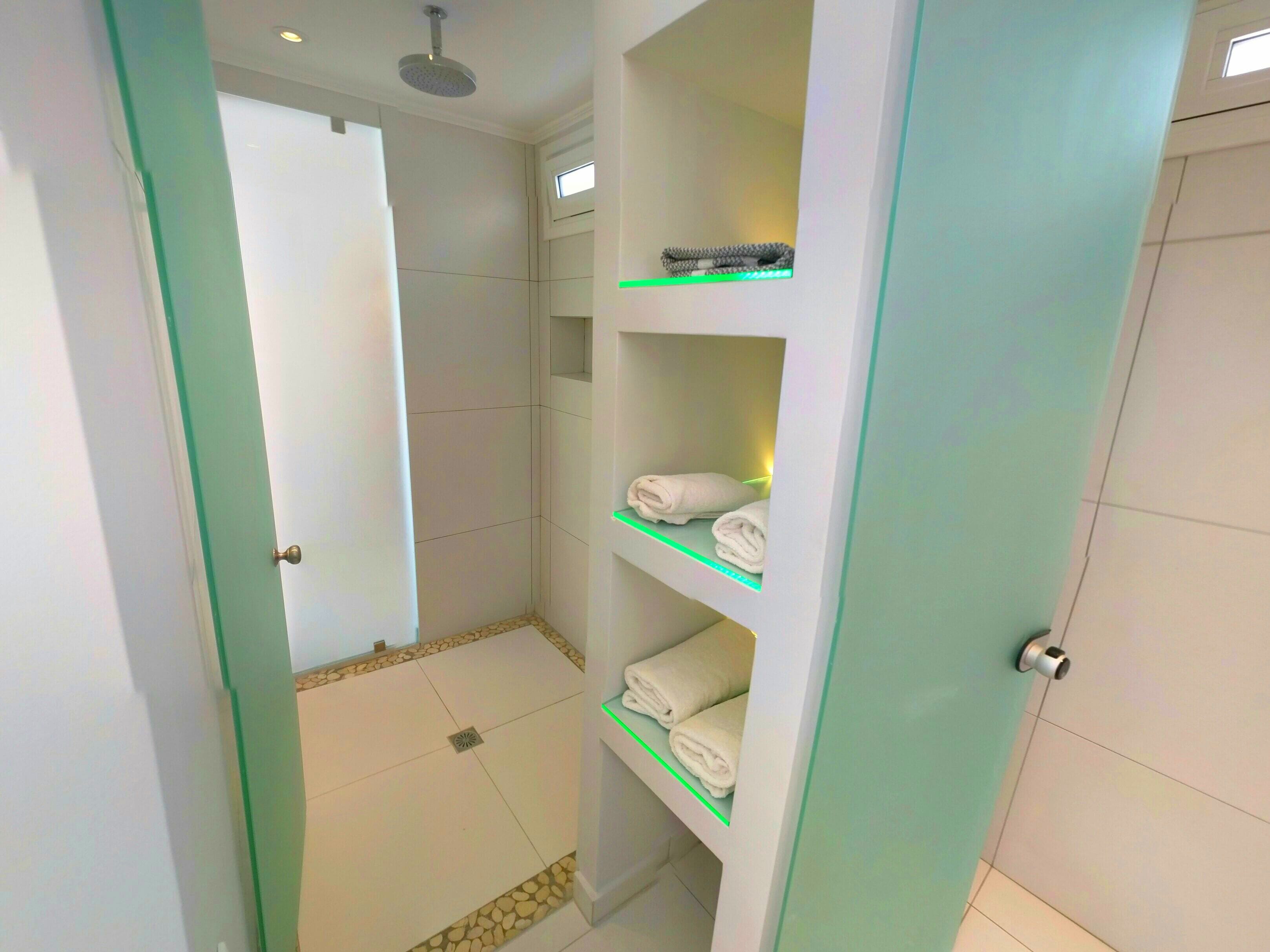 A modern bathroom with a shower, a toilet, and a glass-enclosed vanity with shelves for towels.
