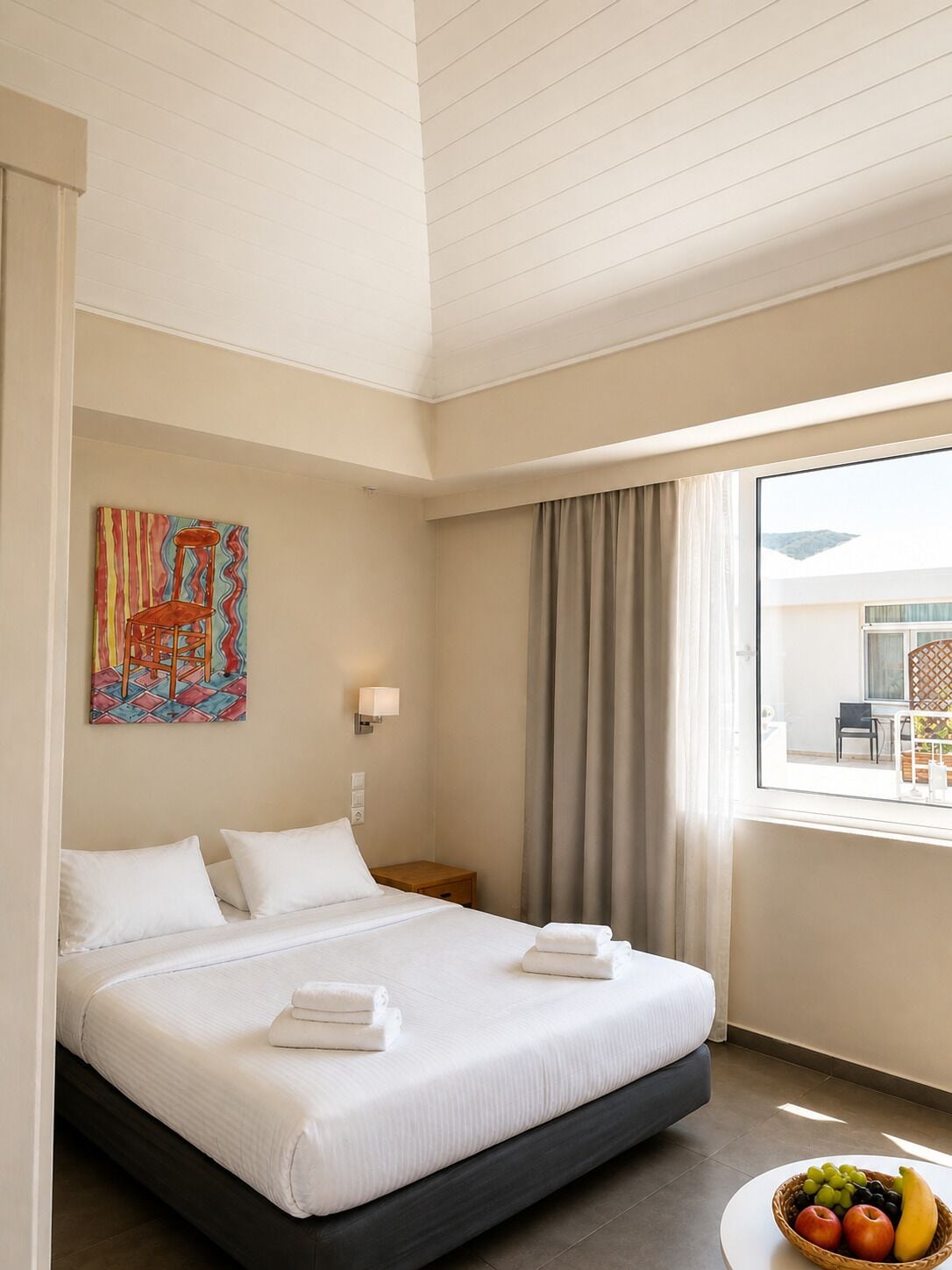 A hotel room with a bed, a painting on the wall, a nightstand, a window with curtains, and a fruit bowl on the floor.