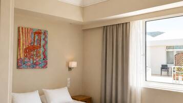 A hotel room with a bed, a painting on the wall, a nightstand, a window with curtains, and a fruit bowl on the floor.