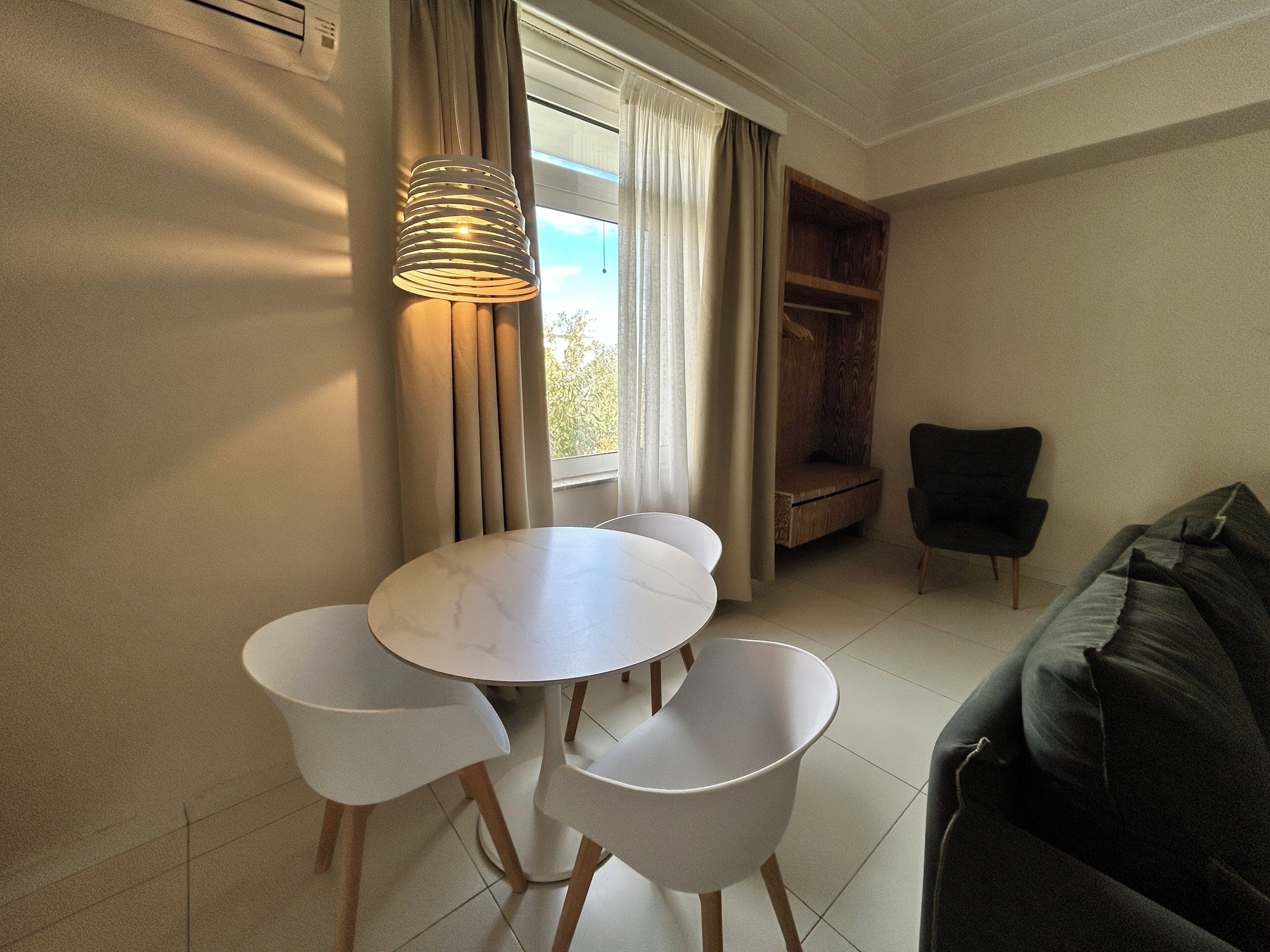 A room with a round table and four chairs, a black armchair, a wooden bench, and a window with curtains.