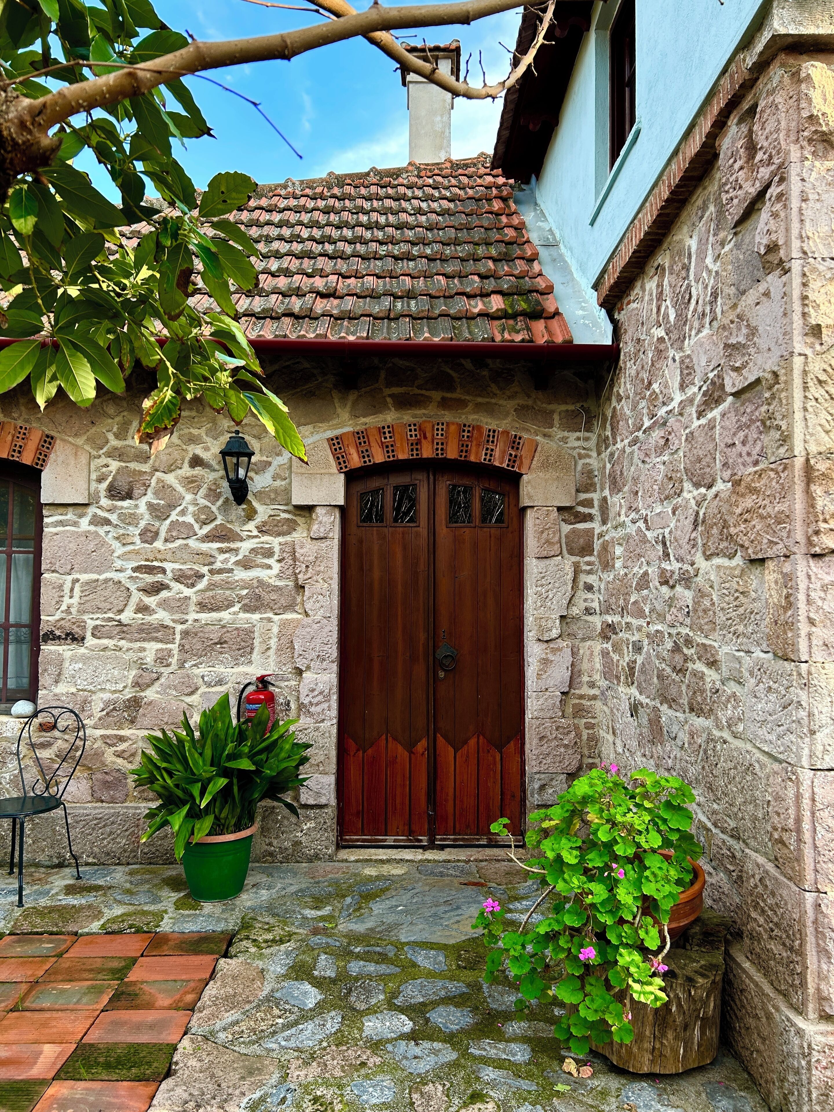 A stone building with a tiled roof, a wooden double door, and a small garden area with potted plants.