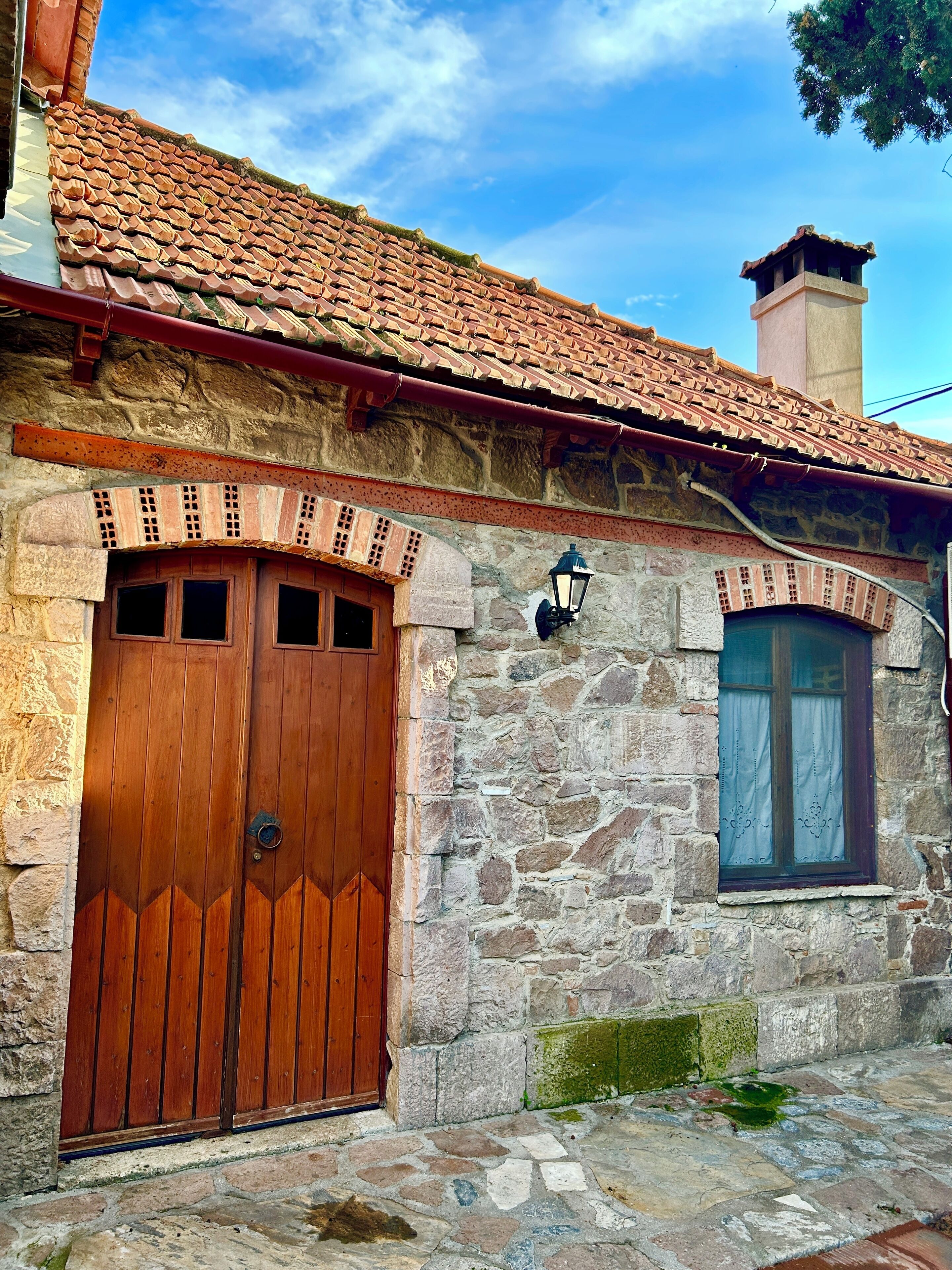 A stone building with a tiled roof, a wooden door, and a window with a curtain.