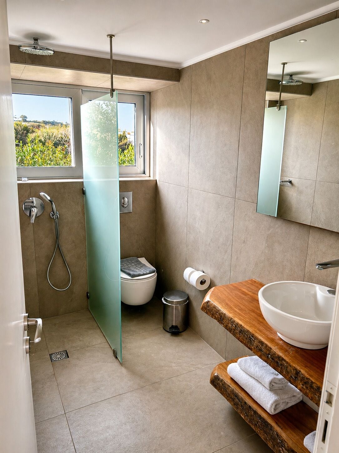 A modern bathroom with a glass shower enclosure, a toilet, a sink with a wooden countertop, and a large mirror.