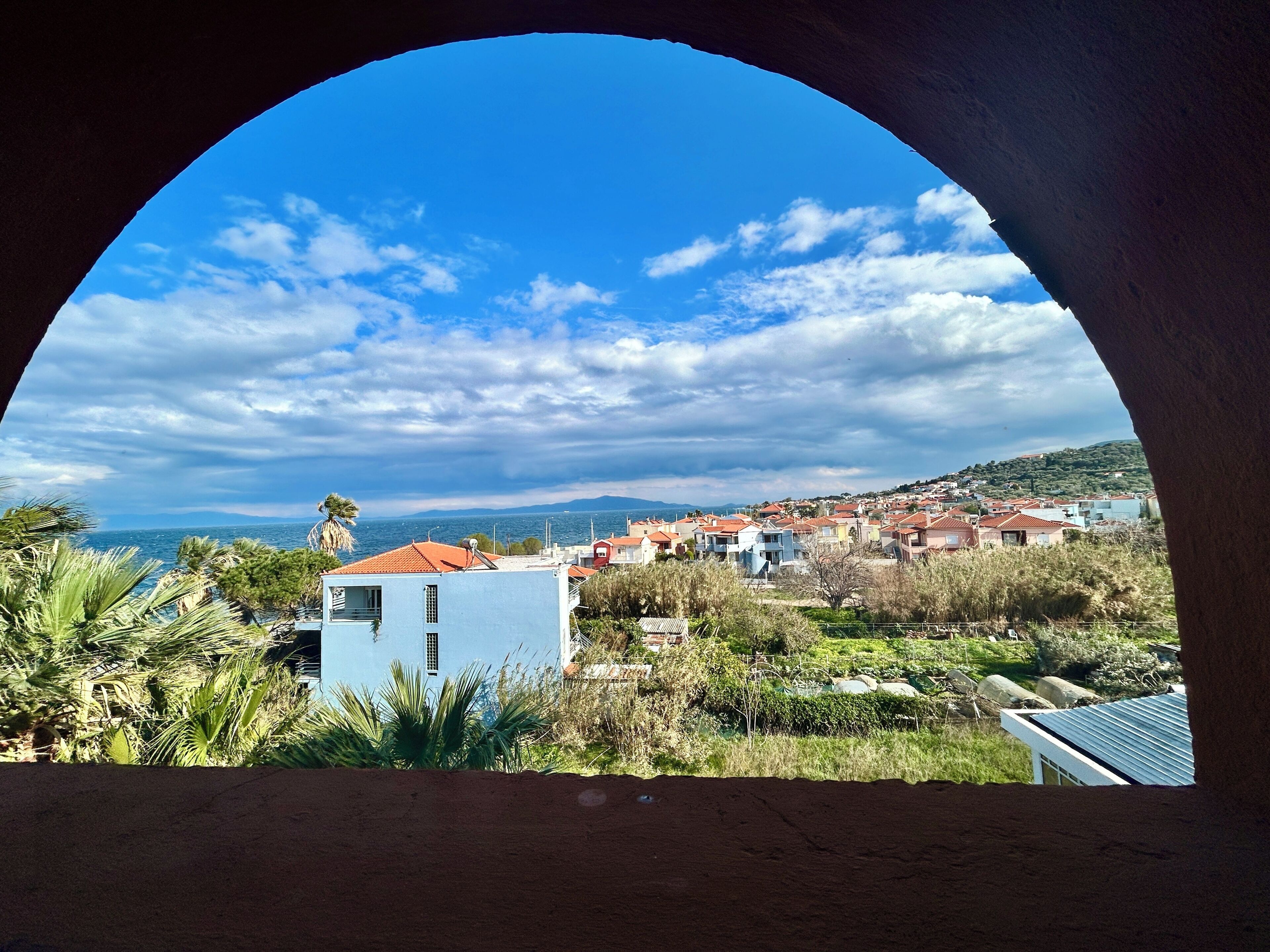 A view through an archway of a coastal town with houses, greenery, and the sea in the background.