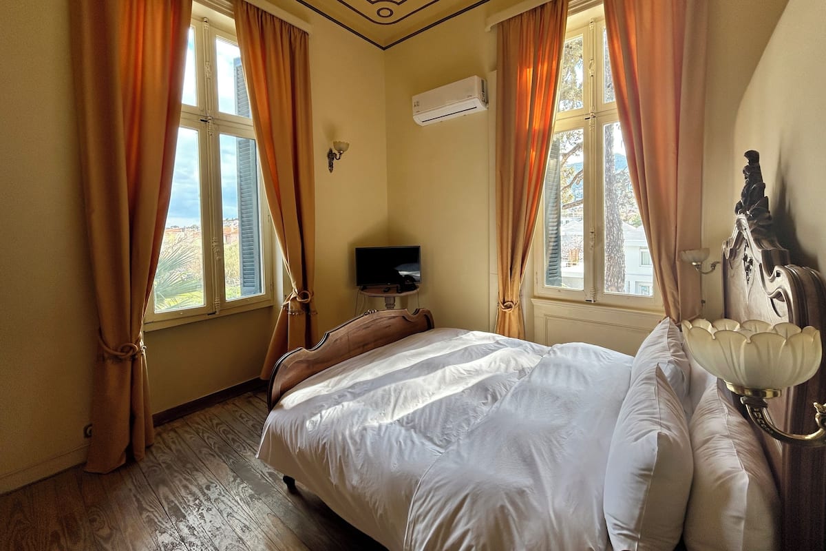 A bedroom with a large bed, two windows with curtains, a flat-screen TV, and an air conditioning unit.
