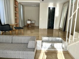 A modern living room with a grey sofa, a black chair, and a small table with two chairs. A staircase with marble steps leads to a dark room.