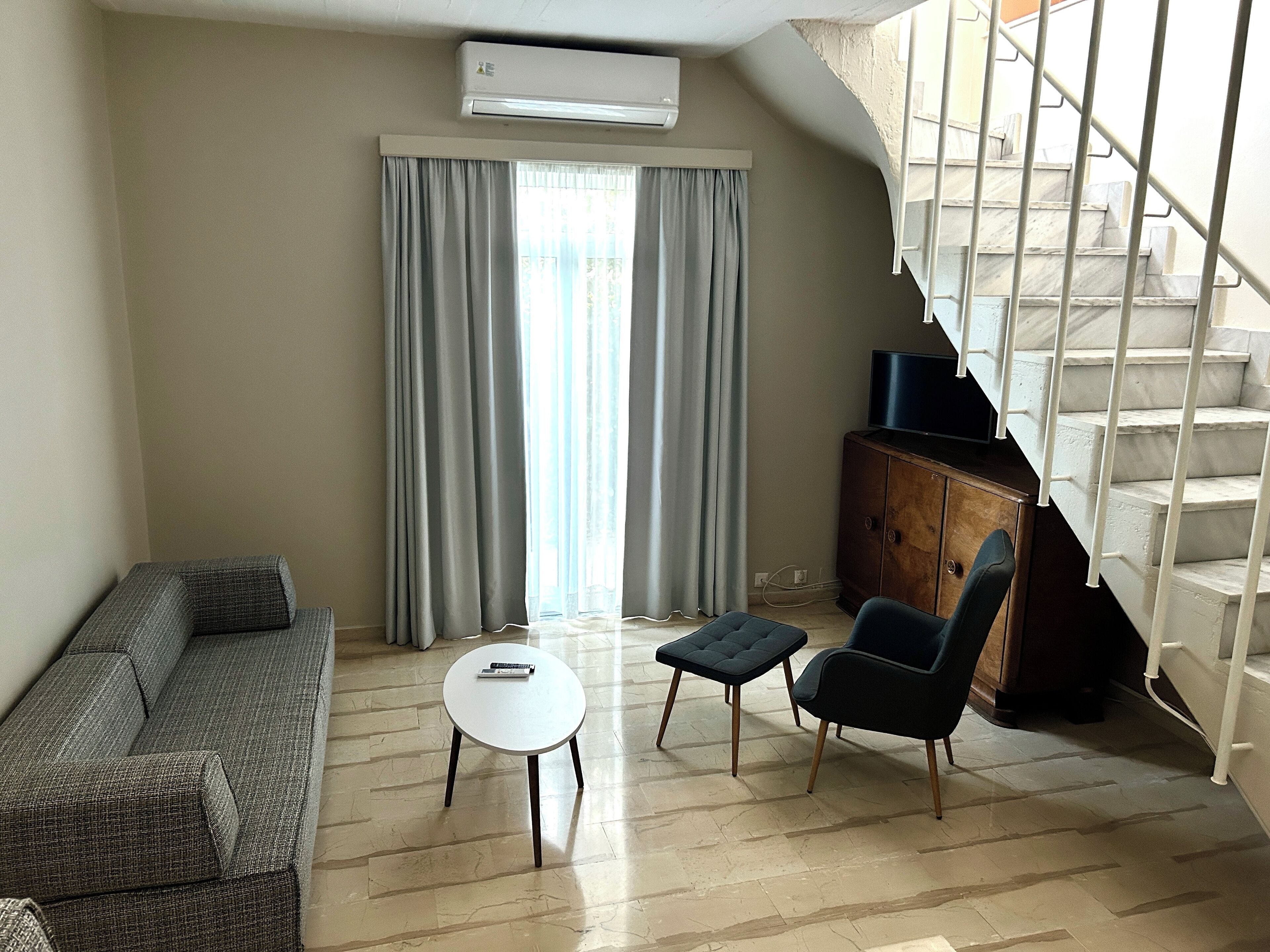 A living room with a sofa, two chairs, a small table, and a TV on a wooden cabinet. A staircase is visible in the background.