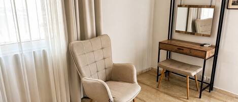 Ein Zimmer mit einem Bett, einem Sessel, einem Schreibtisch, einem Spiegel und einem fensterbepflanzten Fenster.