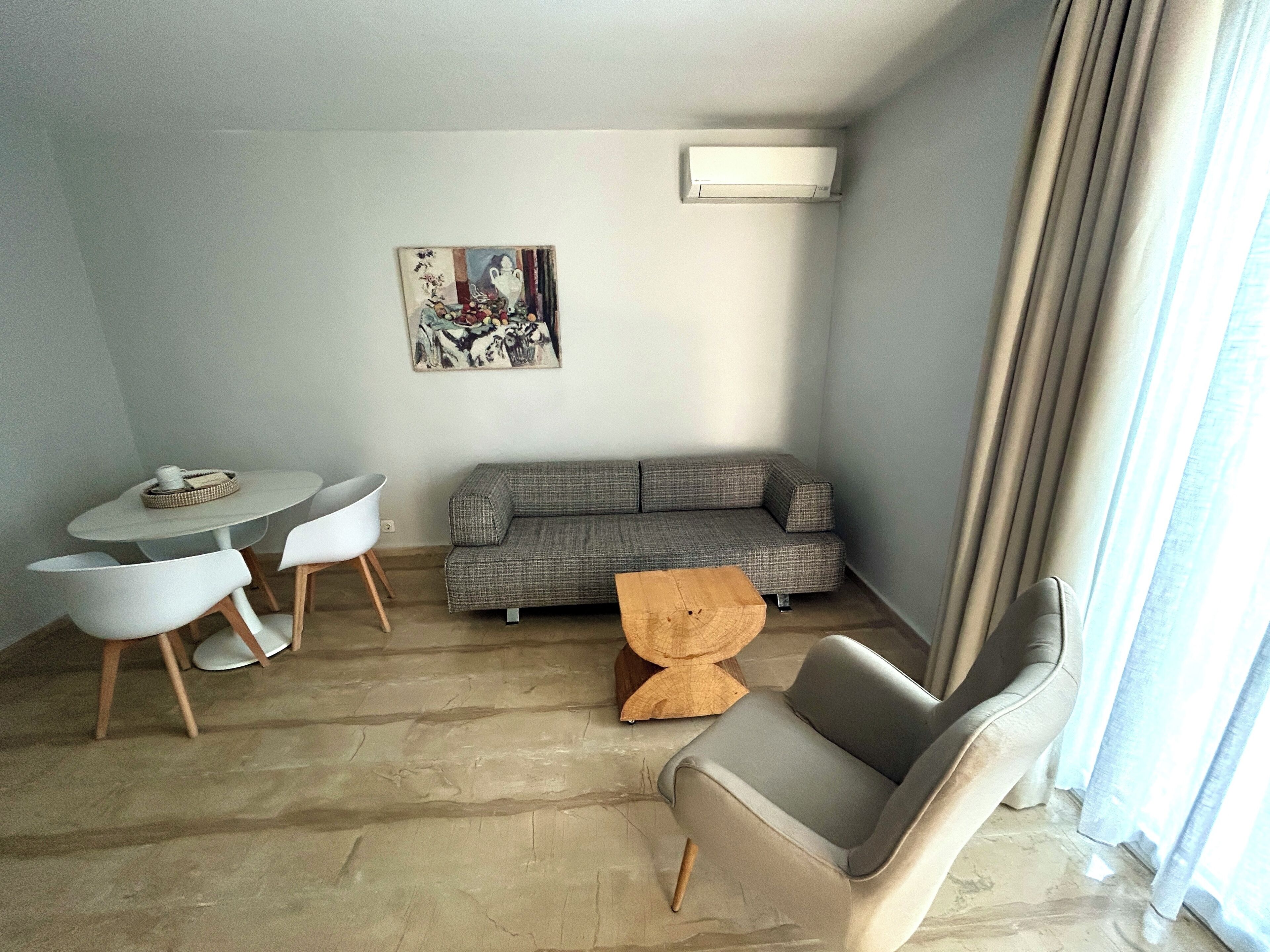 A living room with a grey sofa, a round table with two chairs, a small wooden side table, and a painting on the wall.