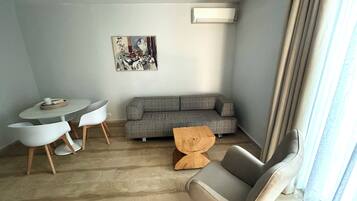 A living room with a grey sofa, a round table with two chairs, a small wooden side table, and a painting on the wall.