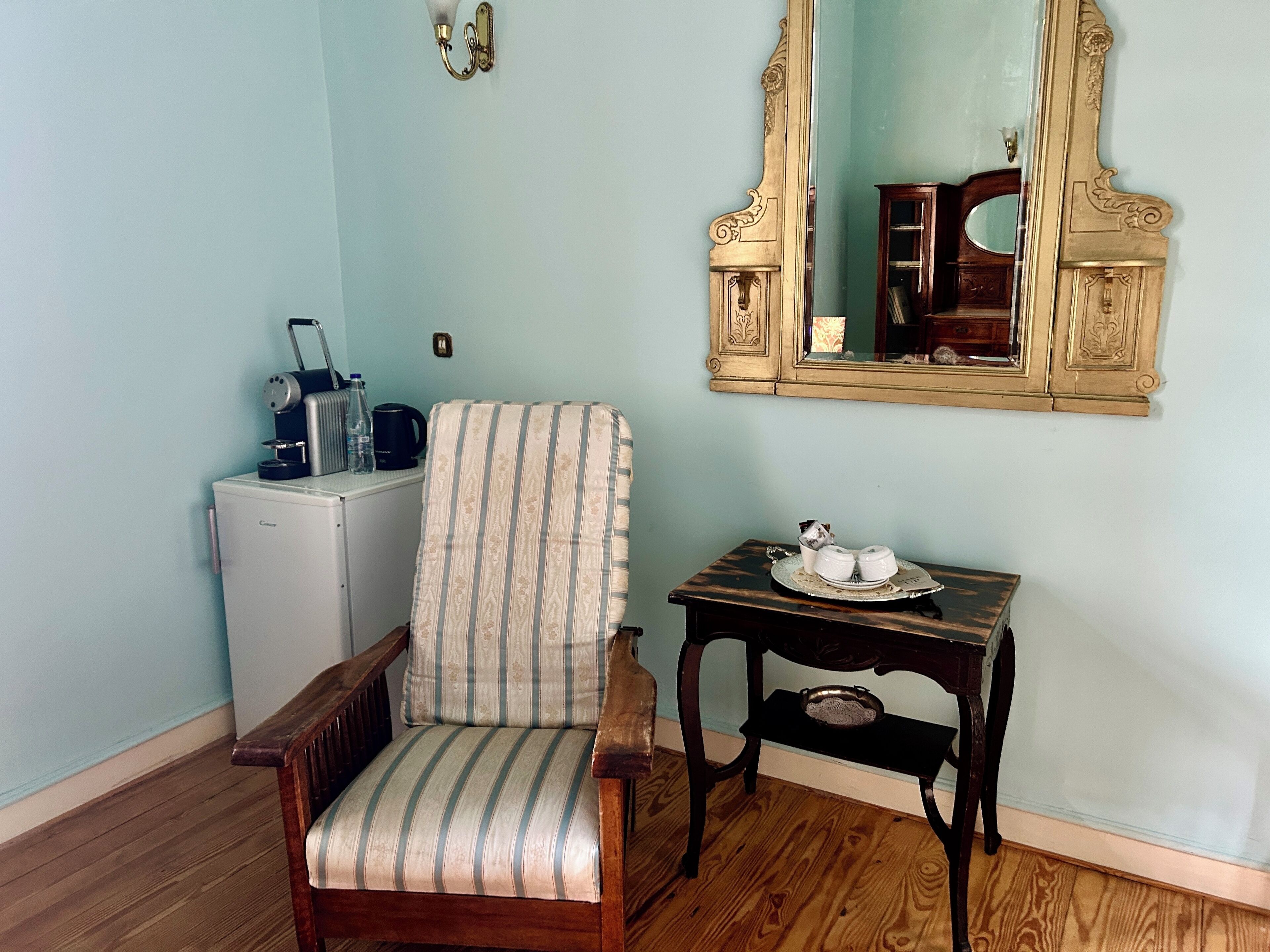 A room with a striped armchair, a small table with a tray, a refrigerator, and a large mirror on the wall.