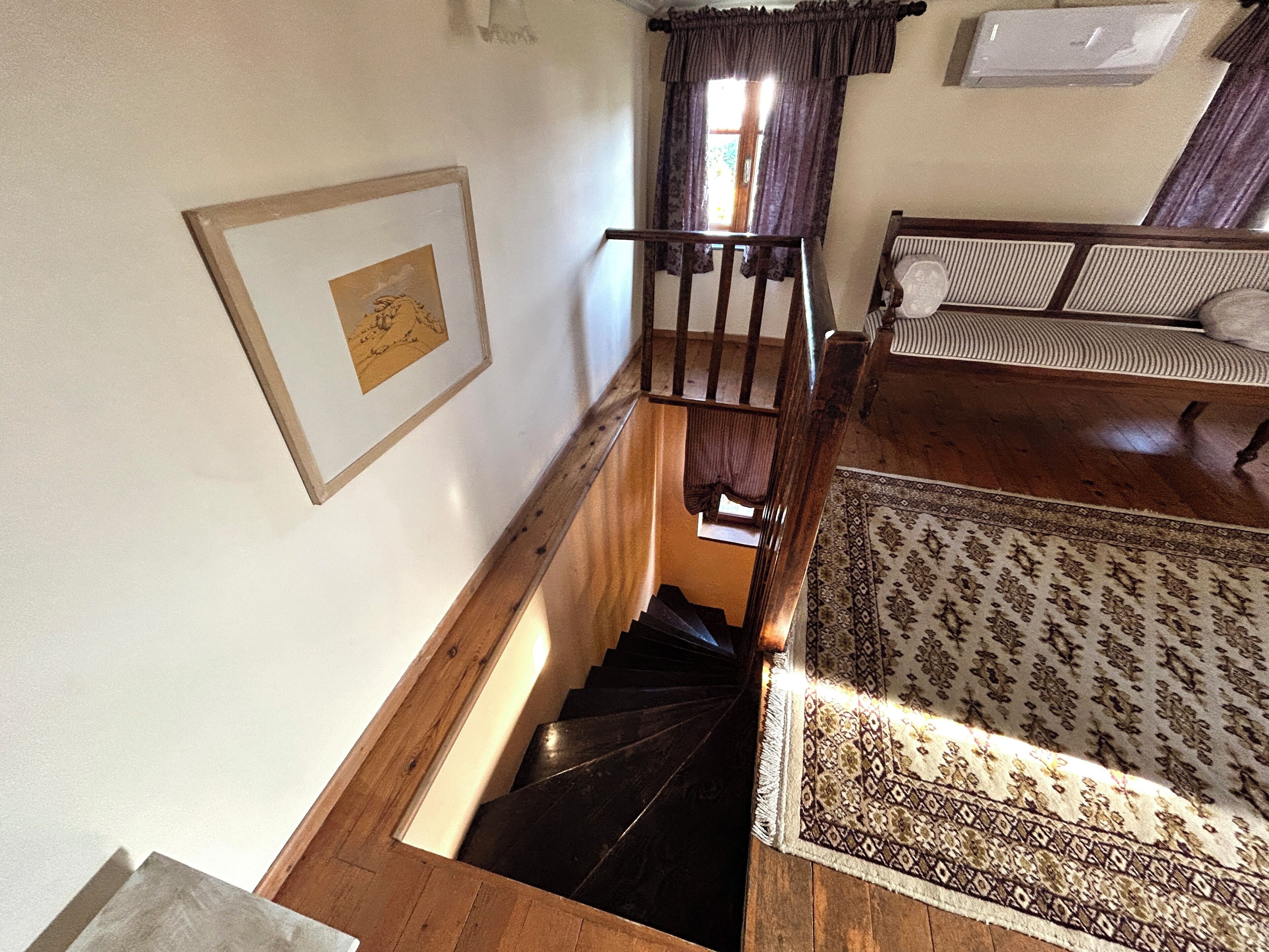 Una escalera conduce a una sala de estar con un tapete estampado, un cuadro enmarcado y una ventana con cortinas.