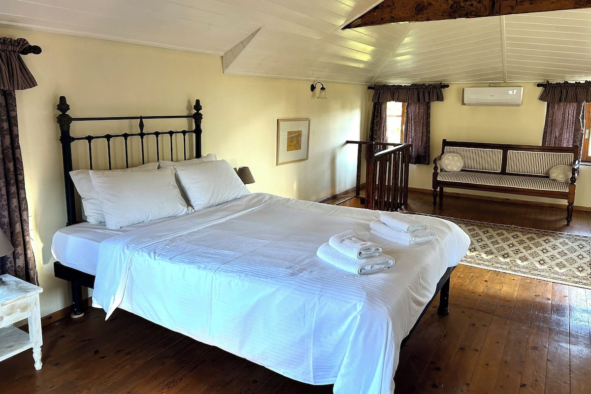 A neatly made bed with white linens and folded towels, a wooden bench, and a patterned rug on a wooden floor.