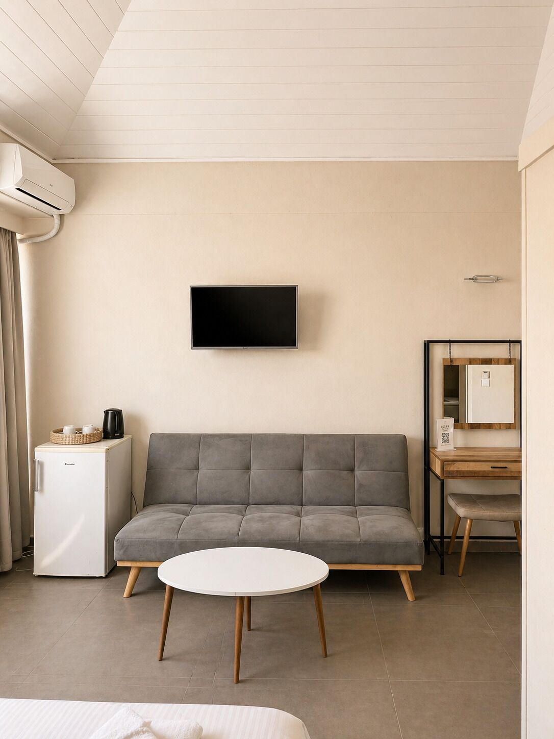 Ein modernes Hotelzimmer mit grauem Sofa, einem kleinen weißen Tisch, einem flachbildfernseher an der Wand, einem kompakten Kühlschrank und einem Holzbüro mit Stuhl.