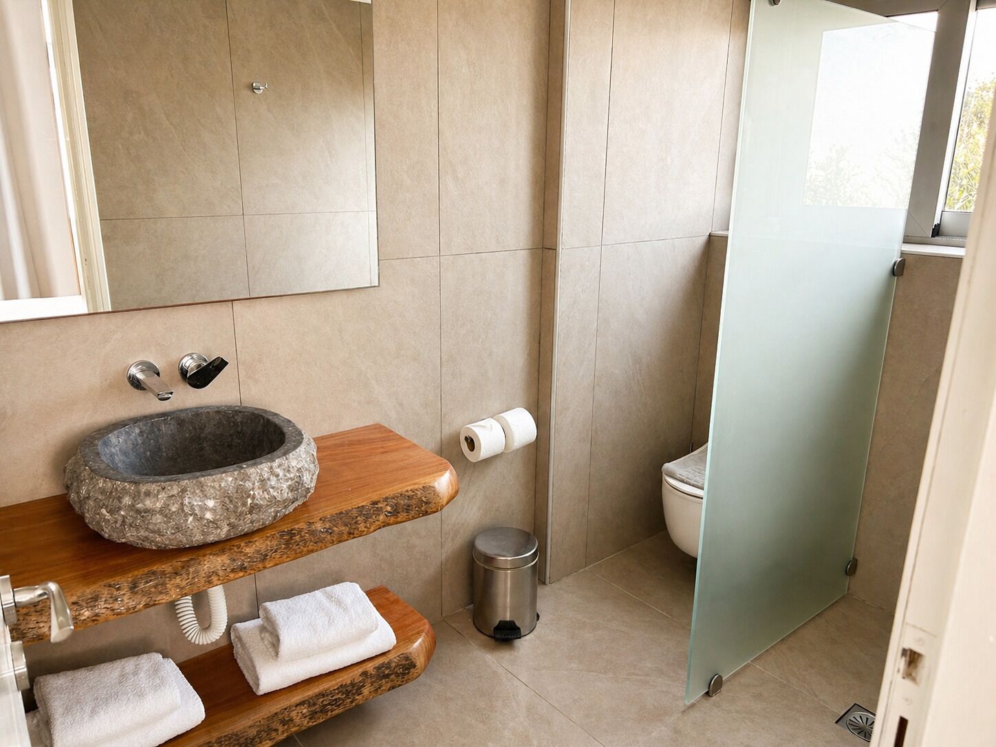 Un baño moderno con un lavabo de piedra, estantes de madera, un inodoro y una puerta de ducha de cristal.