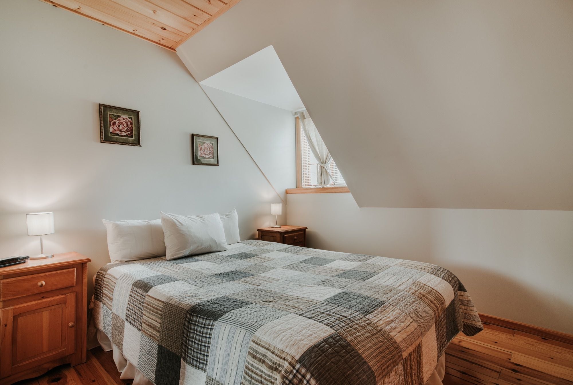 Une chambre avec un plafond en pente, un lit recouvert d’une couverture à carreaux, deux lampes de chevet et deux tableaux encadrés accrochés au mur.