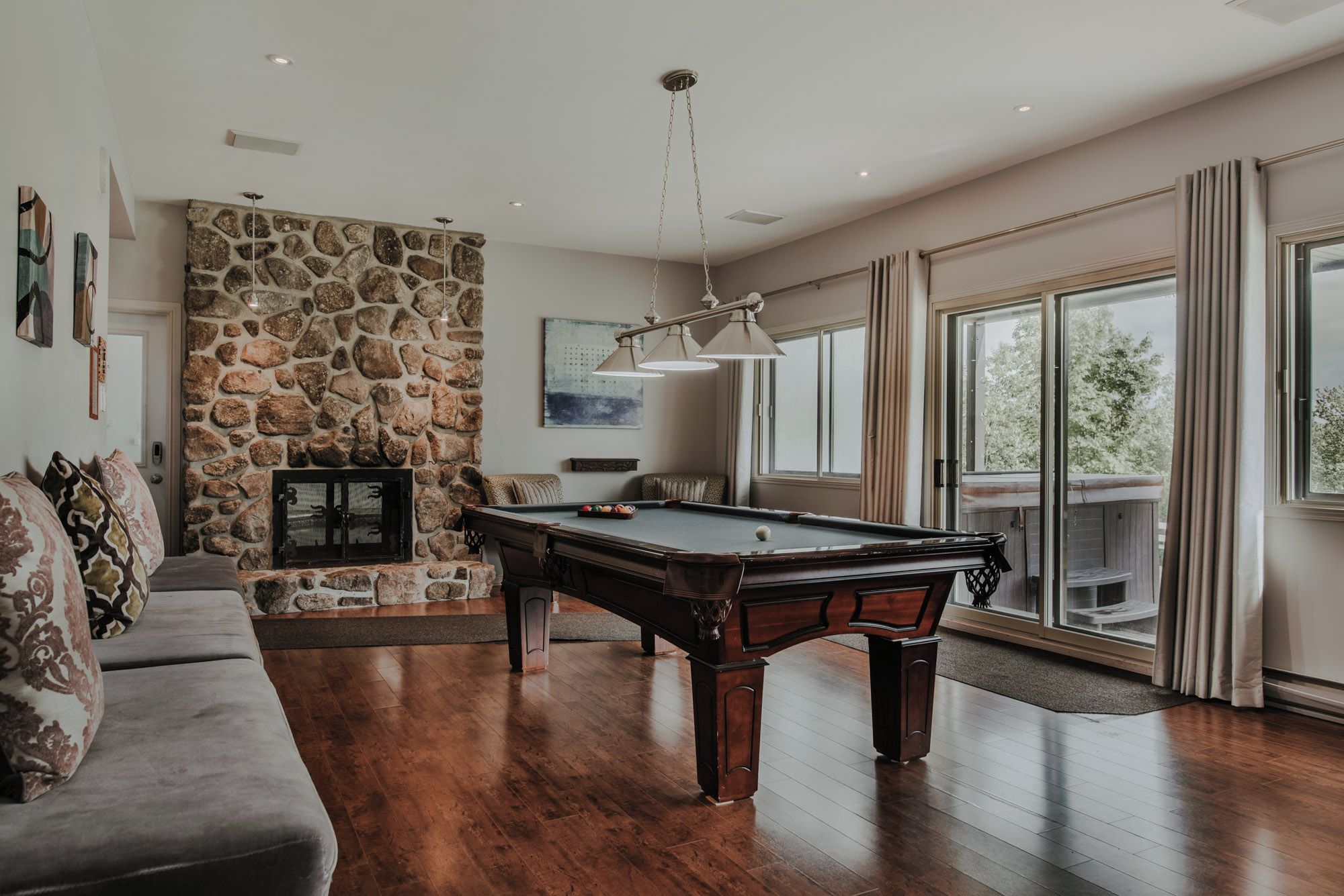 A spacious living room with a pool table, a stone accent wall, and a large window offering a view of the outdoors.