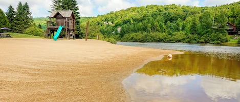 A sandy beach with a wooden play structure and a small house in the background.