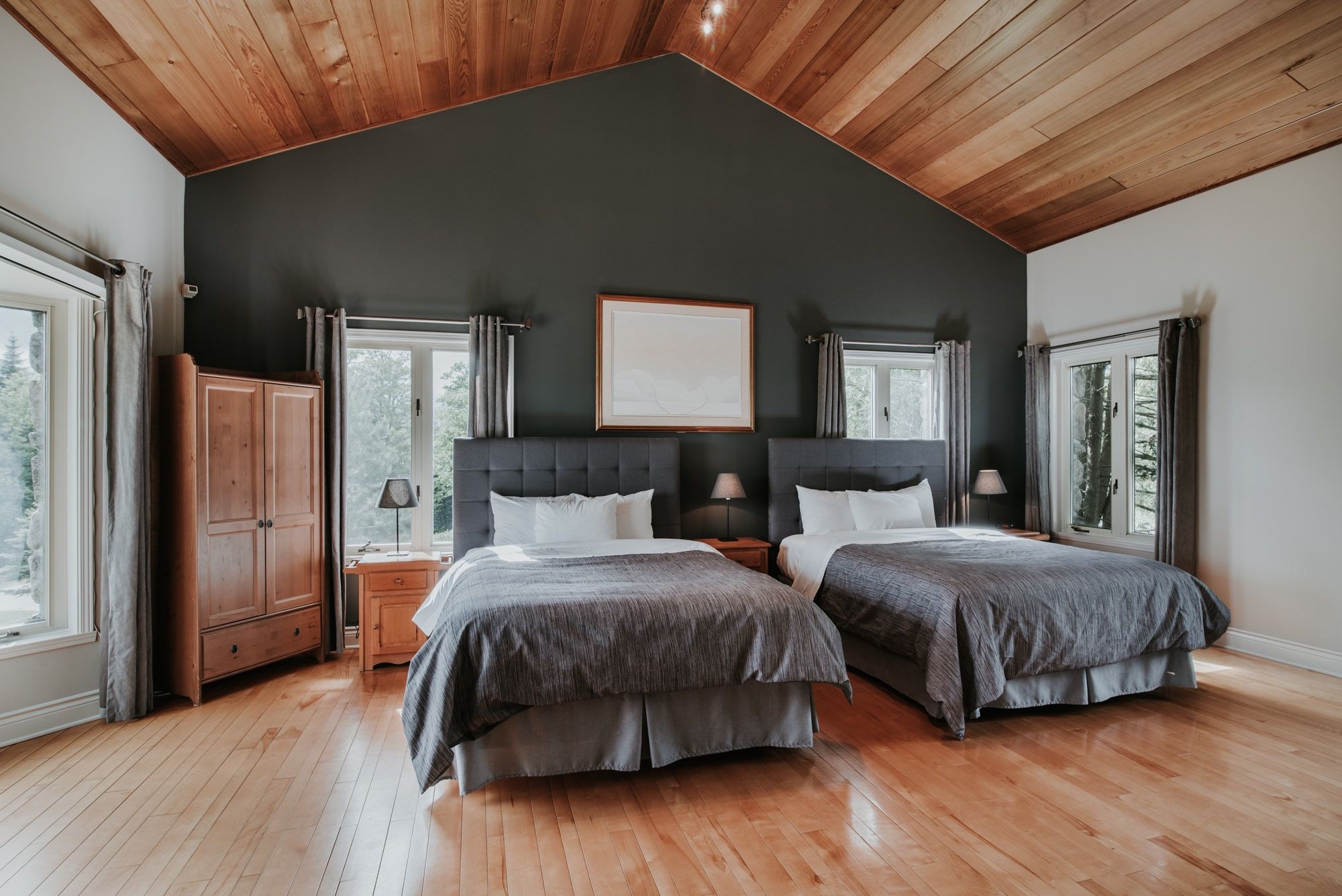 A bedroom with two beds, wooden flooring, a dark ceiling, and large windows.