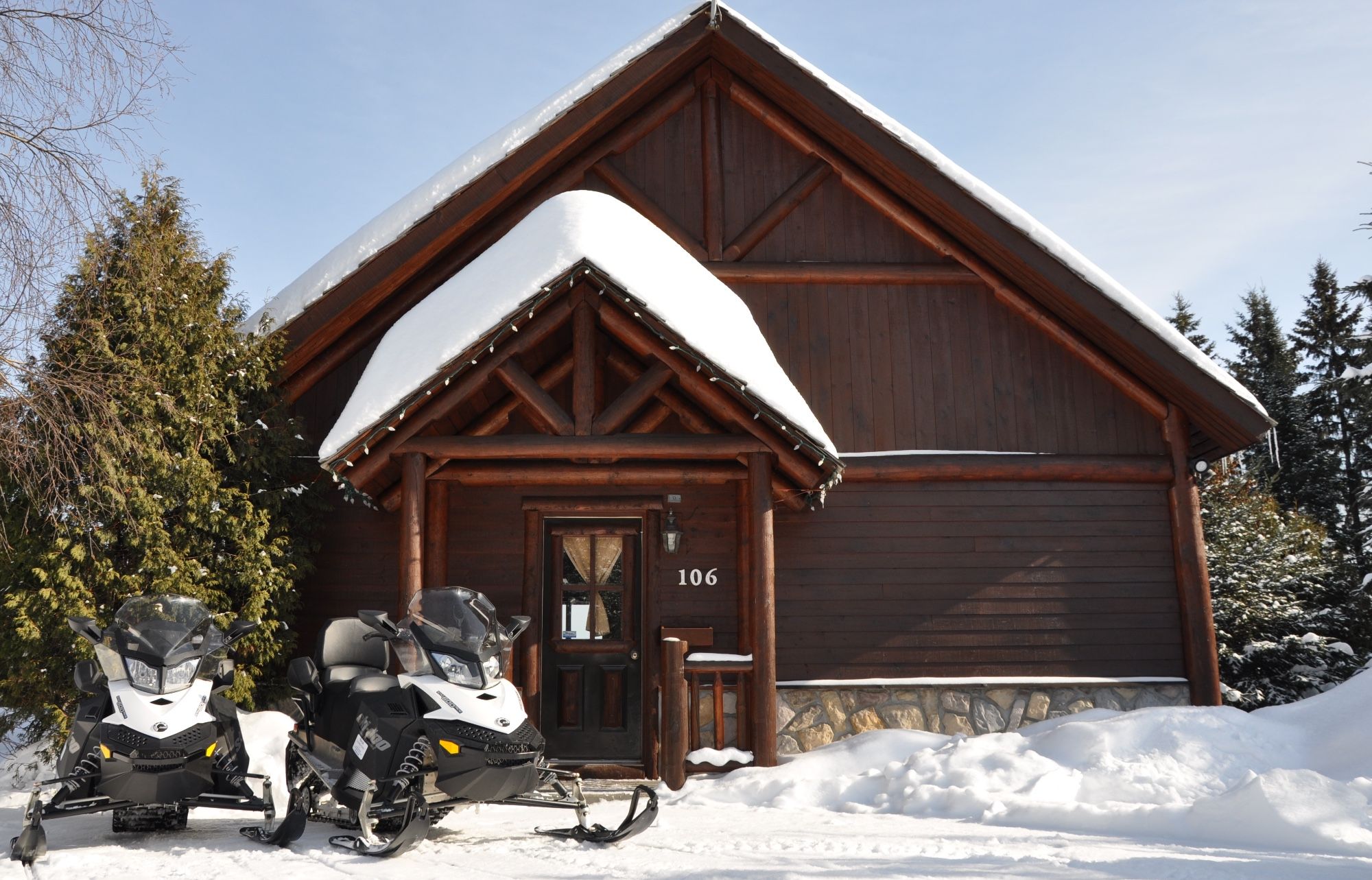 A wooden lodge with a snow-covered roof, a visible number '106' on the door, and a snowmobile parked in front.