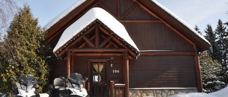 A wooden lodge with a snow-covered roof, a visible number '106' on the door, and a snowmobile parked in front.