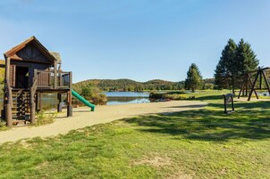 A playground with a wooden playhouse, a slide, and a swing set near a lake.
