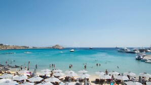 A beach with clear turquoise water, numerous umbrellas, and boats. - The George Hotel (Mykonos)