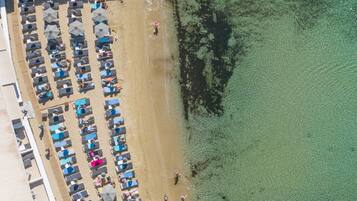 Vista aérea de una playa con filas de tumbonas y sombrillas, agua clara y poco profunda, y algunas personas caminando por la orilla.