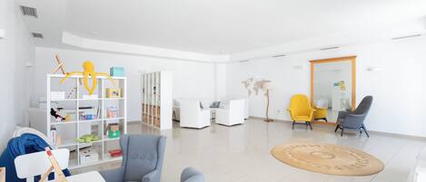 A spacious living room with a white sofa, a grey armchair, and a yellow armchair. There is a white bookshelf filled with various items and a large world map on the wall.