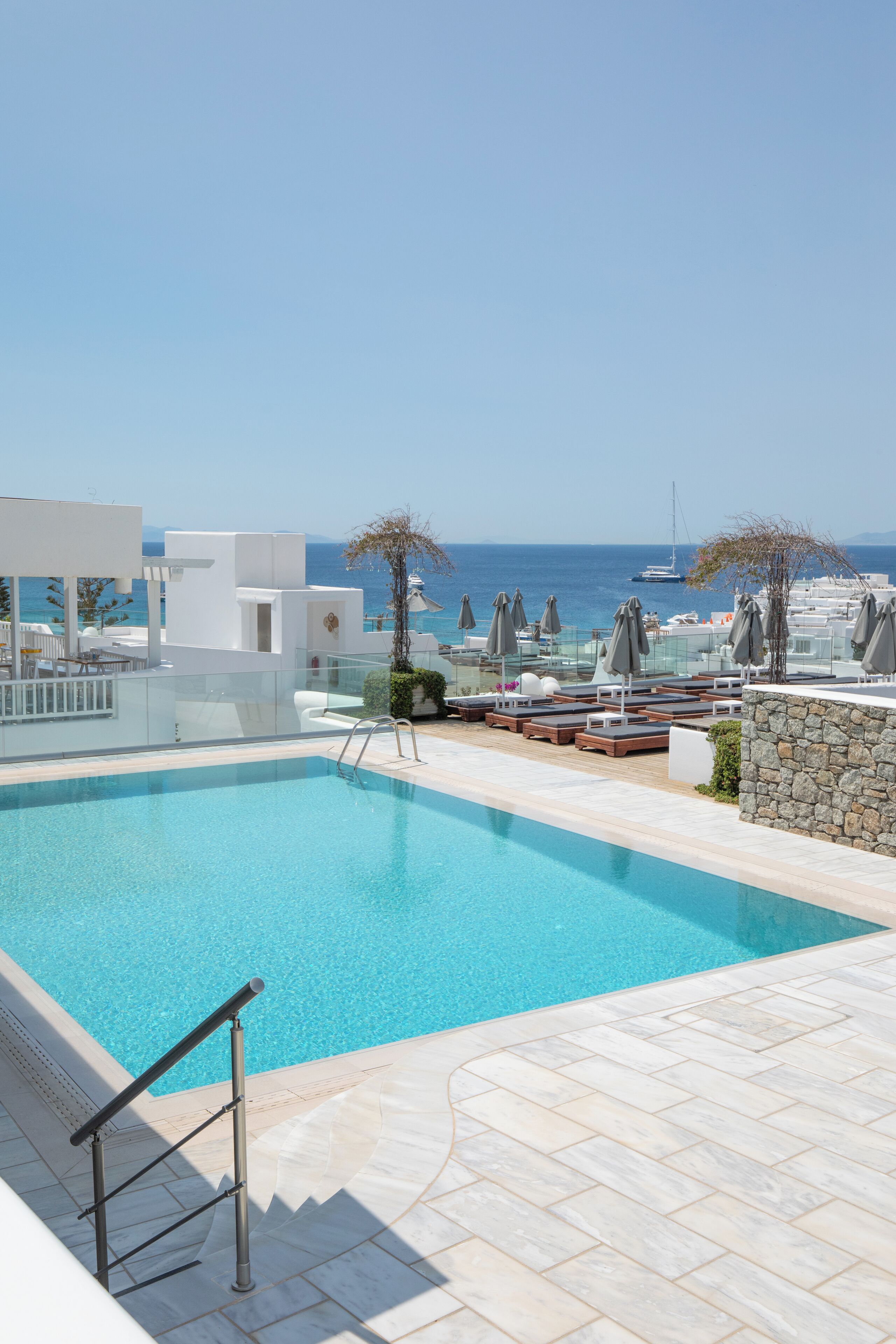 Piscina con sillones de descanso y sombrillas, un muro de piedra y vistas al mar y a las embarcaciones.