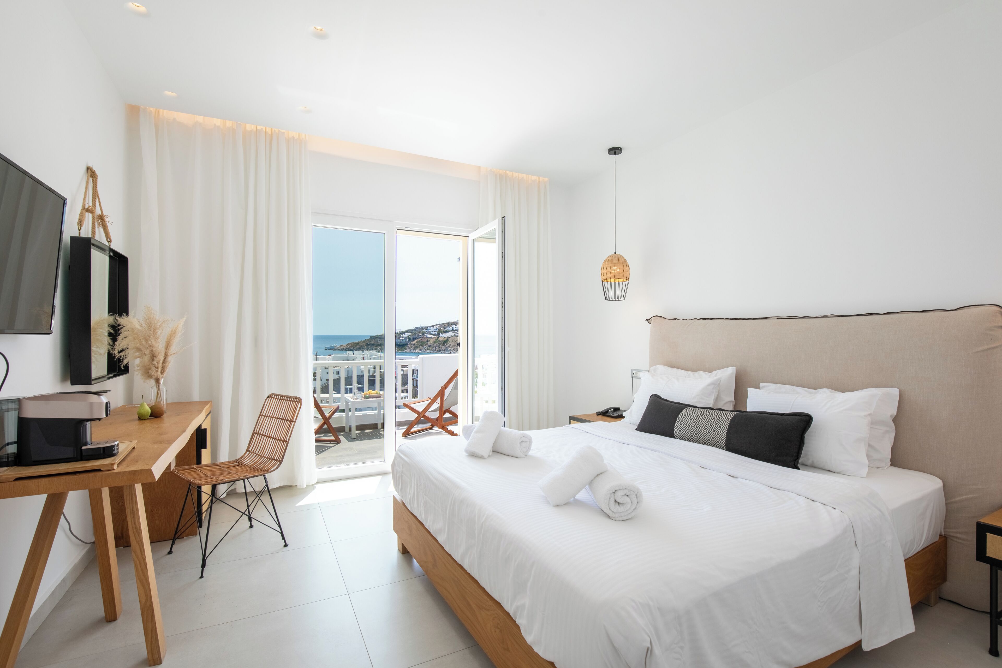 A modern hotel room with a large bed, a desk with a computer, and a view of the sea through the balcony doors.