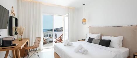 A modern hotel room with a large bed, a desk with a computer, and a view of the sea through the balcony doors.