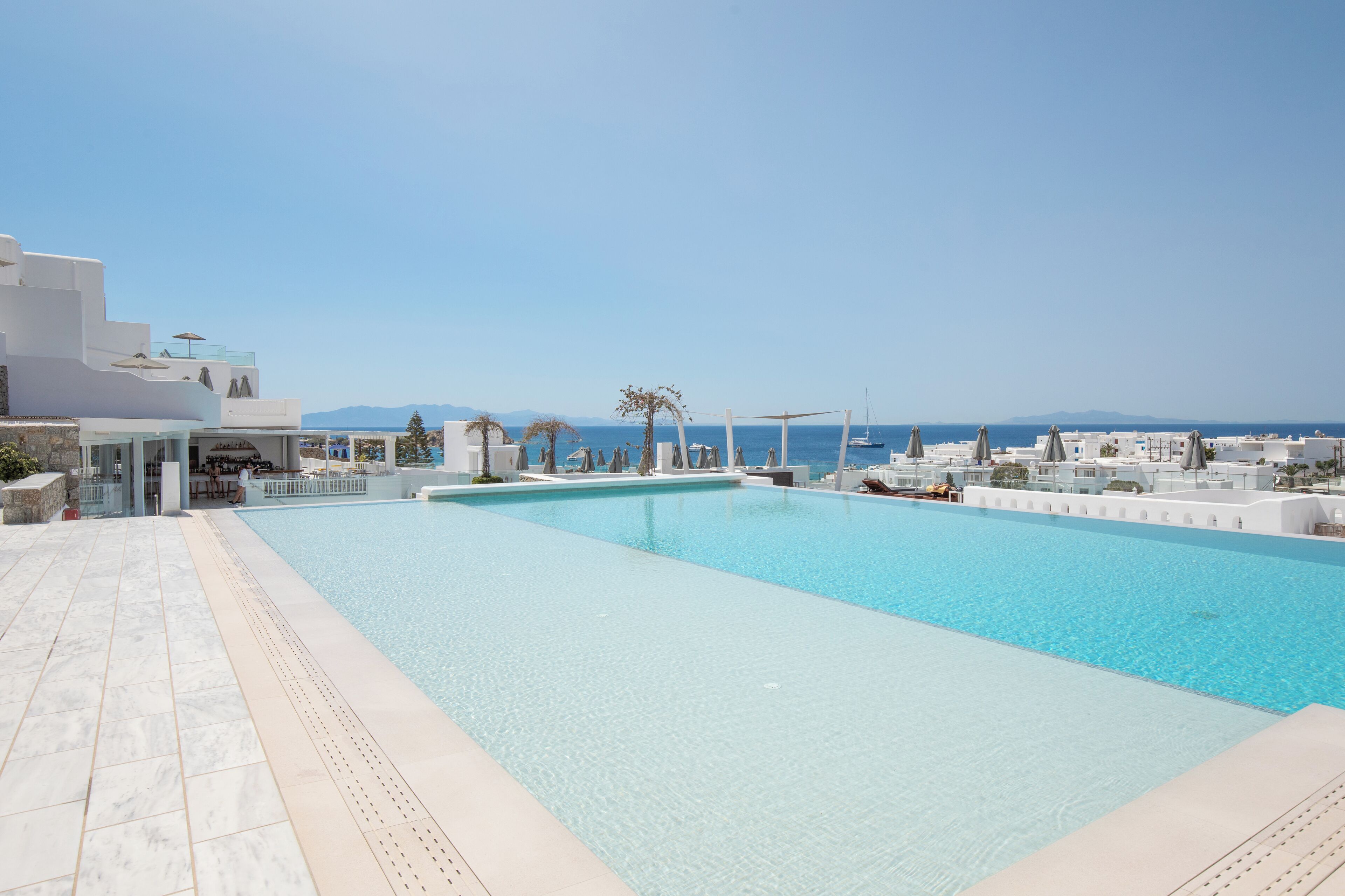 A rooftop swimming pool with clear blue water, surrounded by white buildings and a view of the sea.