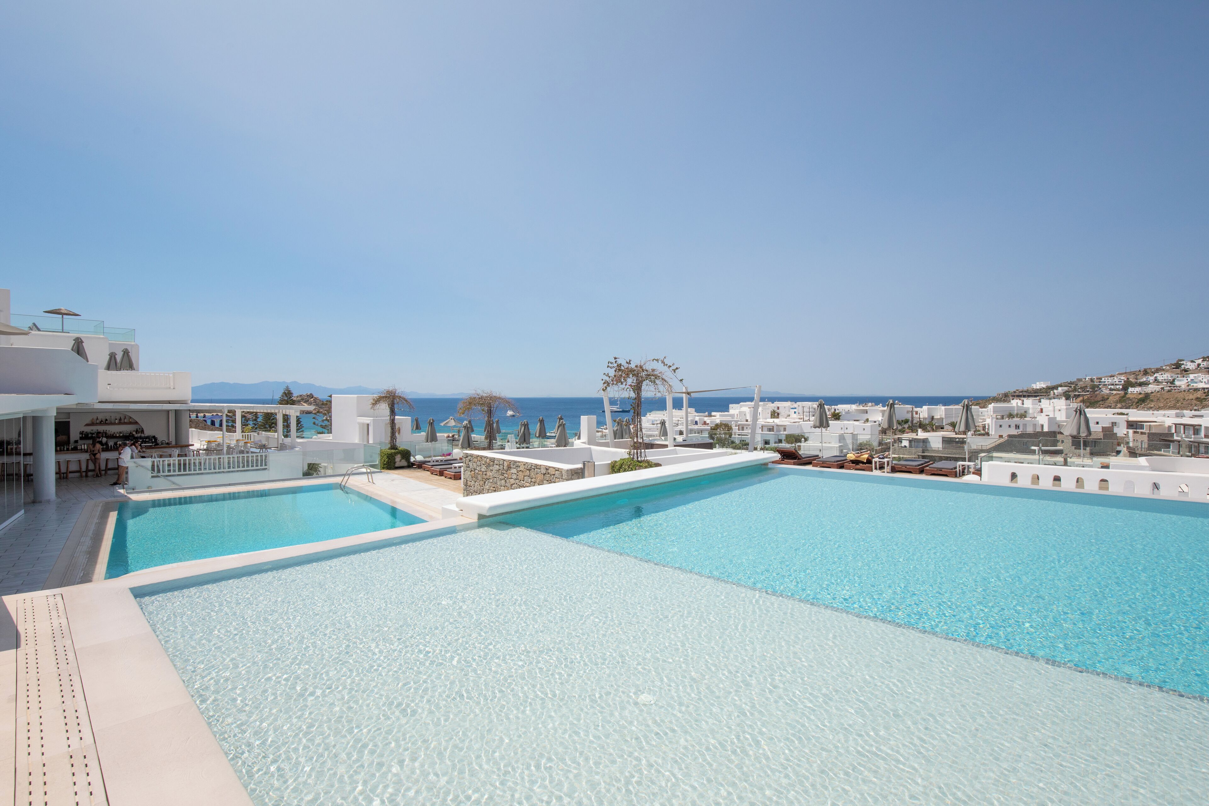 A large outdoor swimming pool with clear blue water, surrounded by white buildings and a view of the sea.