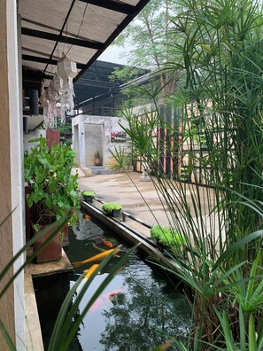 Un bassin à carpes koï, avec des plantes aquatiques et un petit pont, entouré de verdure et bordé d’un bâtiment sur lequel est apposée une enseigne.