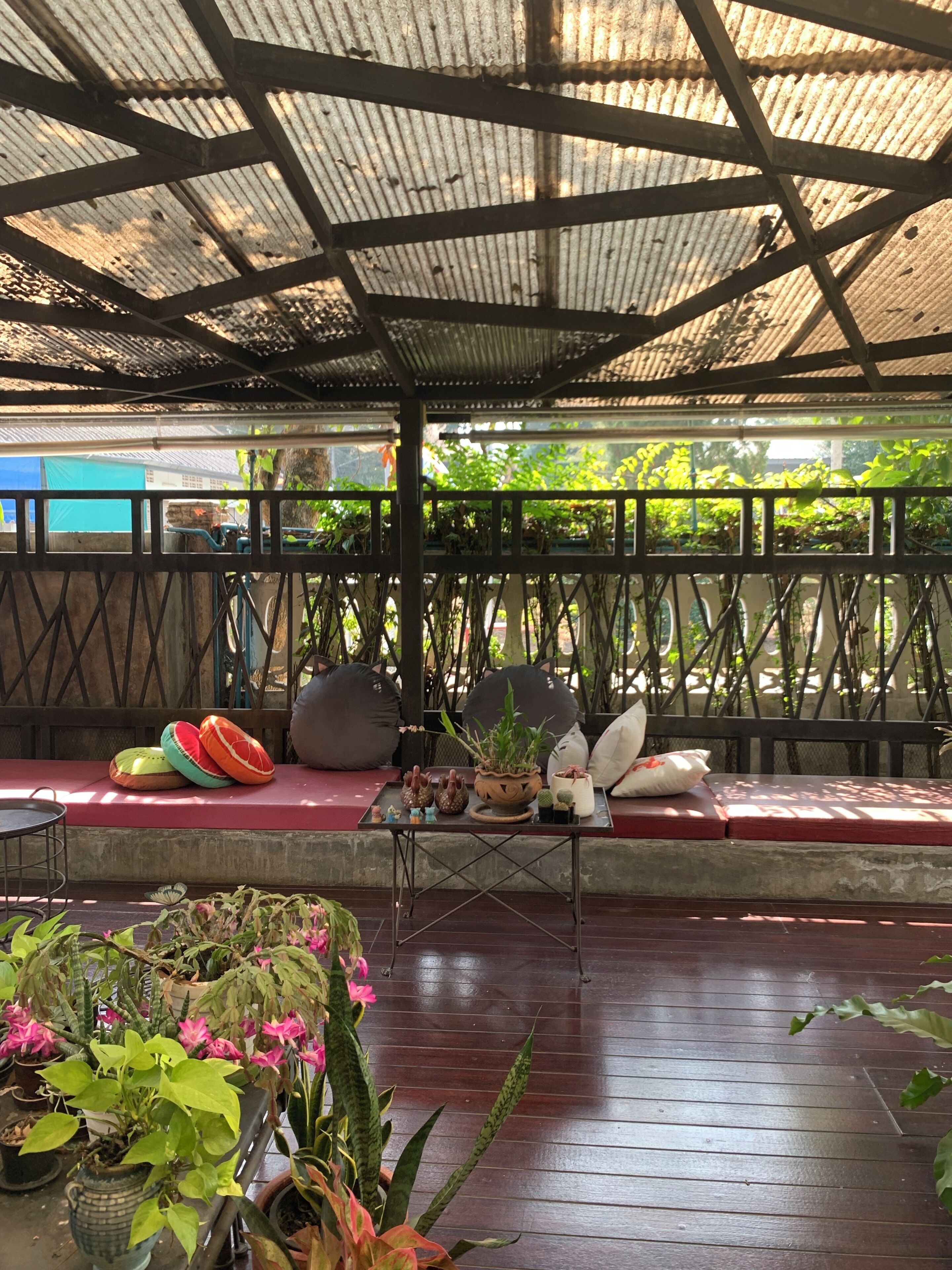A covered outdoor seating area with a wooden floor, cushioned benches, and potted plants.