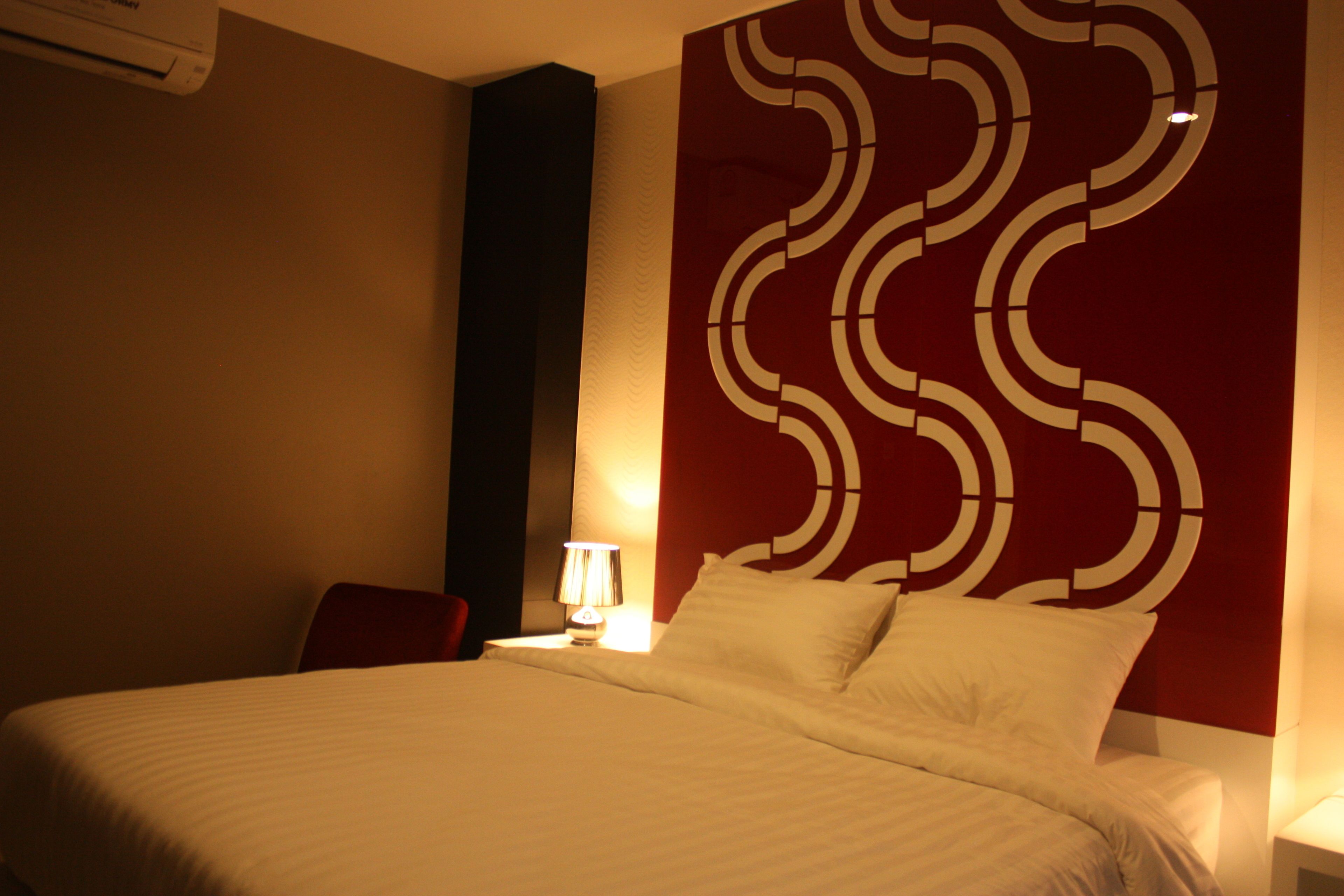 A hotel room with a bed, a red accent wall, and a lamp on a bedside table.