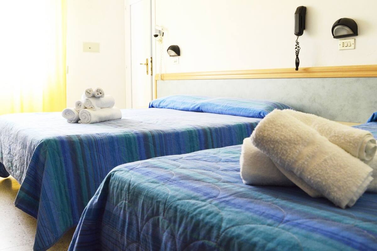 A hotel room with two beds, each with a blue and white striped bedspread. There are rolled towels on the beds, and a phone on the wall.