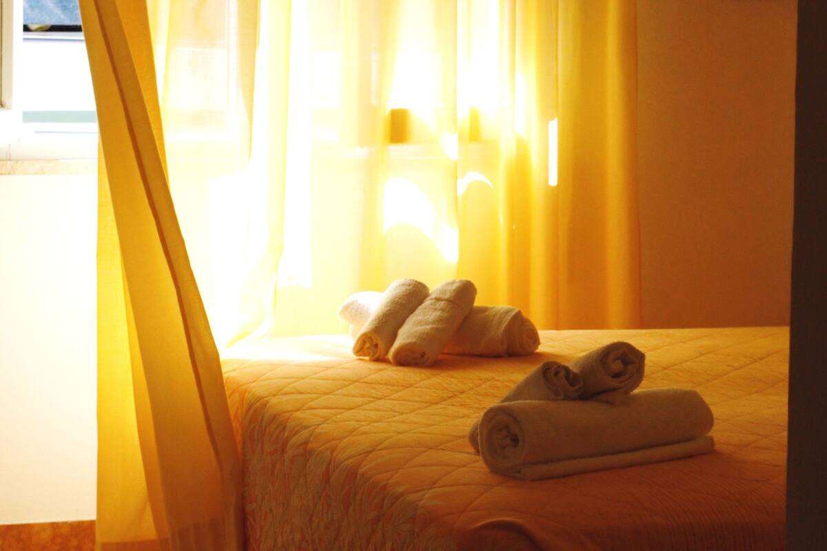 A hotel room with a bed, yellow curtains, and rolled towels.
