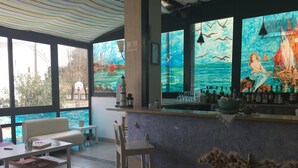 A bar area with a mural of a mermaid and sailboats, a seating area with a white sofa and table, and a view of the outside through large windows. - Hotel Lux (Cesenatico)