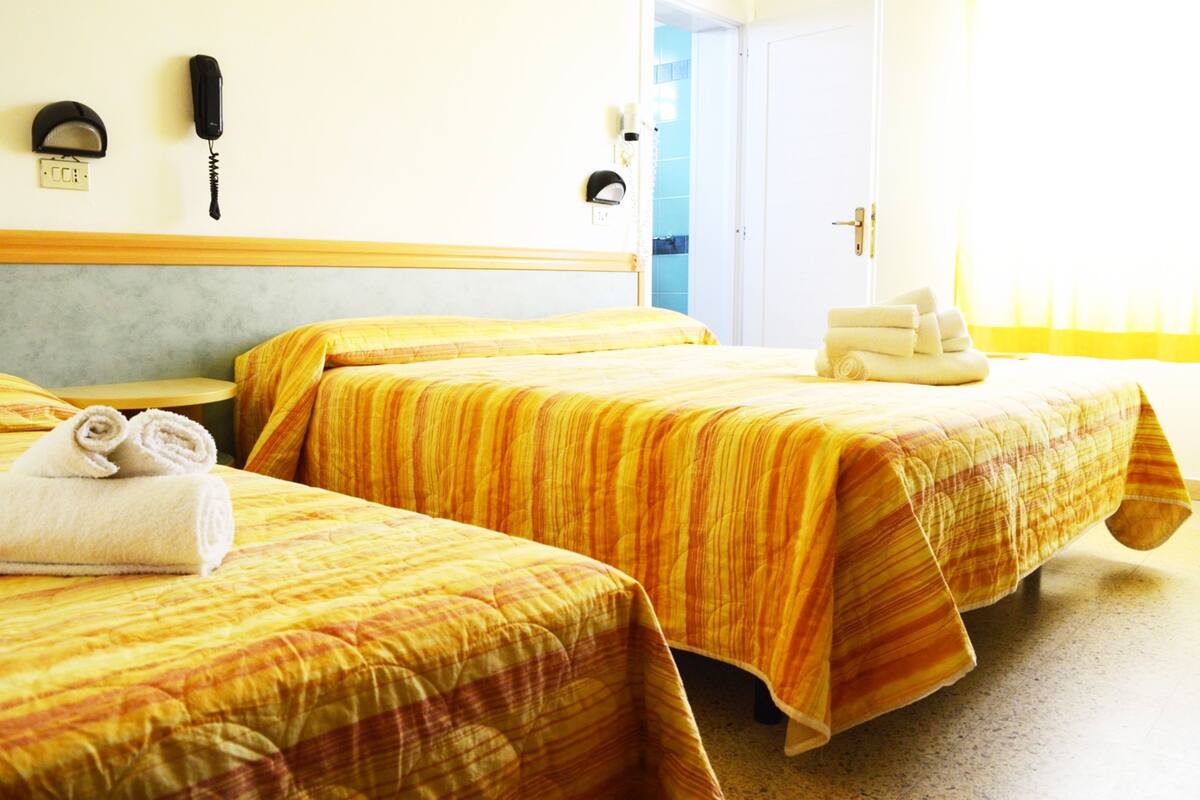 A hotel room with two beds, each with a striped bedspread and rolled towels. There is a telephone and a wall socket on the wall.