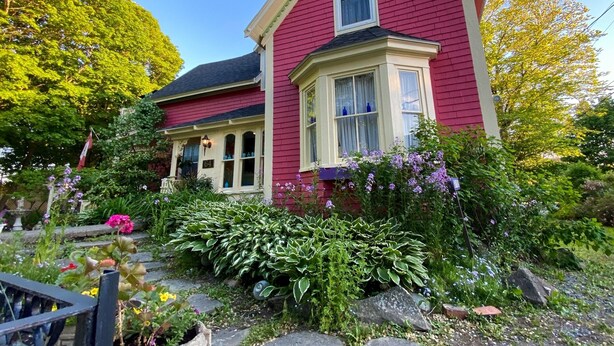 Una casa roja con un ventanal saliente y un porche, rodeada de un jardín con diversas plantas y flores.