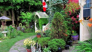 Un jardín con un sendero de piedra que lleva a un porche blanco. Se observan diversas macetas con plantas y flores, y es visible una bandera canadiense.