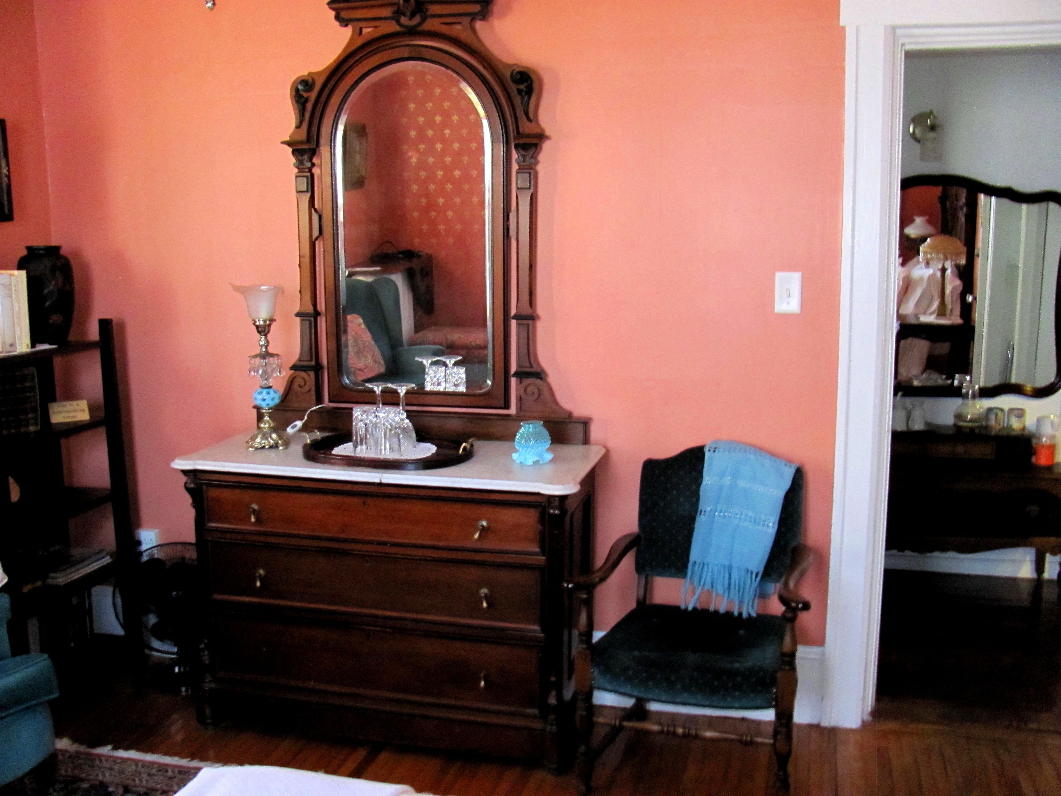 A room with a wooden dresser, a mirror, a chair, and a shelf with books.
