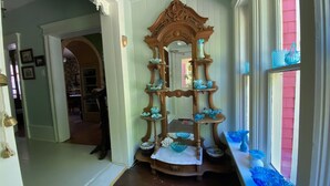 Un pasillo con un expositor de madera que sostiene objetos de vidrio azules, una puerta que da a otra habitación y una ventana con contraventanas rosadas.