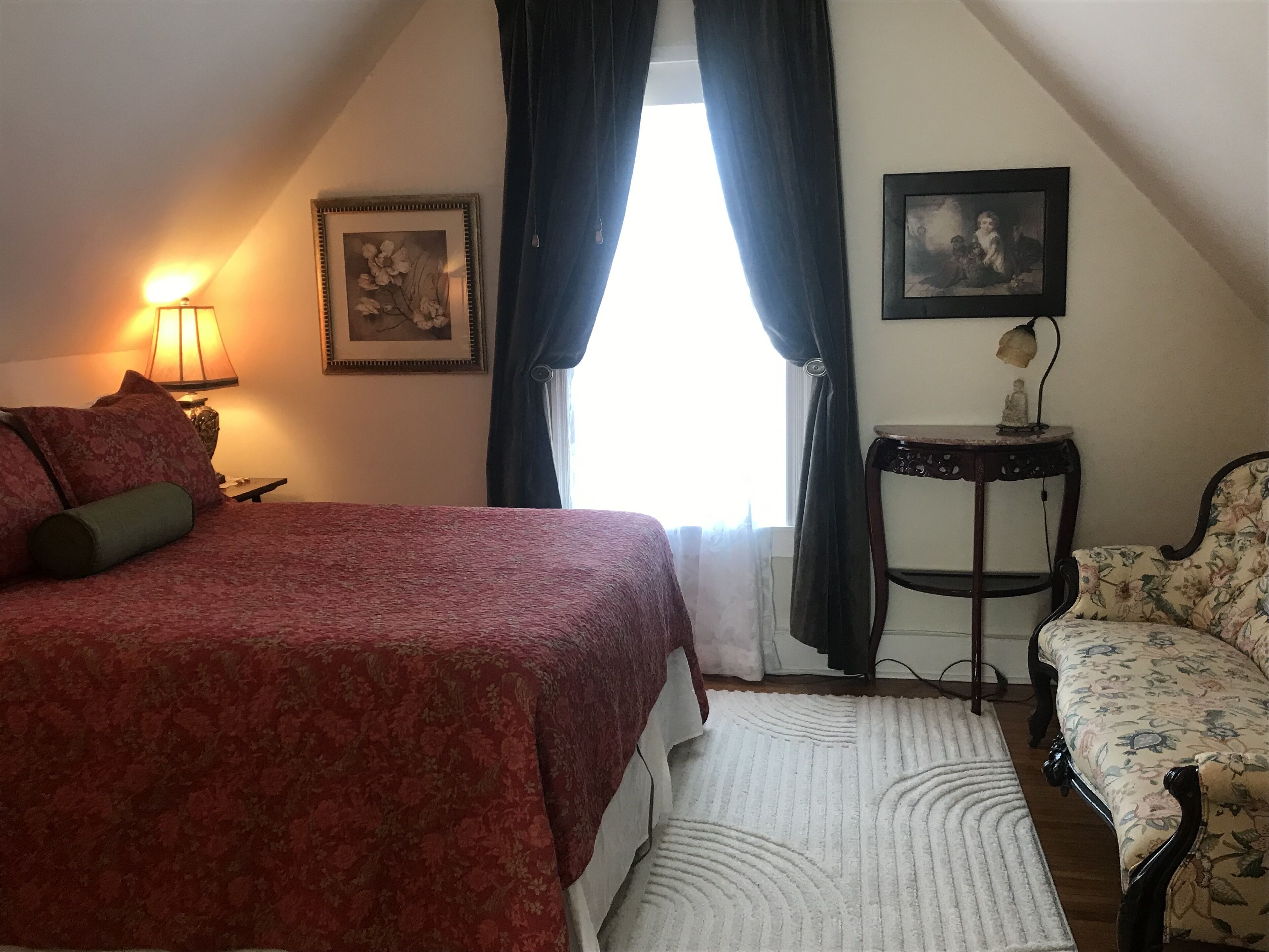 Un dormitorio con cama, una silla, una mesa y dos cuadros en la pared.