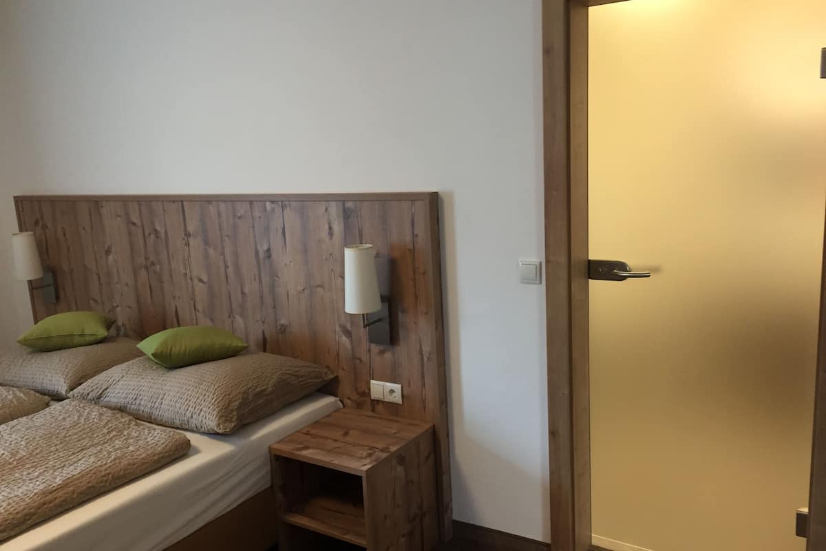 Un dormitorio con cabecera de madera, dos mesitas de noche, una cama con ropa de cama color beige y almohadas verdes, y una lámpara de pared.