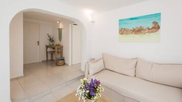 A living room with a beige sofa, a coffee table with flowers, a painting on the wall, and a door leading to another room.