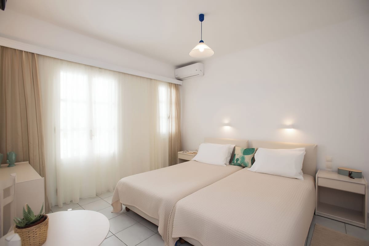 A hotel room with two beds, a bedside table, a lamp, a plant, and a window with curtains.