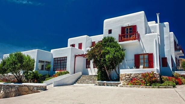 Un bâtiment blanc avec des volets rouges et un balcon, entouré de verdure et surplombé d’un ciel bleu clair.